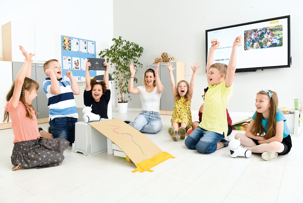 Skupina dětí a učitelka se radují, když hrají s robotem Photon. Děti sedí na zemi a zvedají ruce, některé se usmívají a jiné se smějí. V pozadí je tabule a různé vzdělávací materiály.