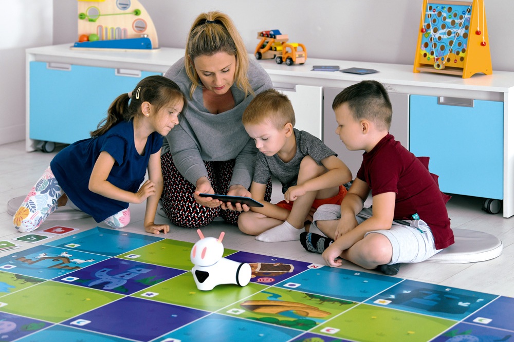 Skupina dětí sedí kolem stolu s robotem a interaktivní hračkou, která reaguje na jejich pohyby a pokyny.
